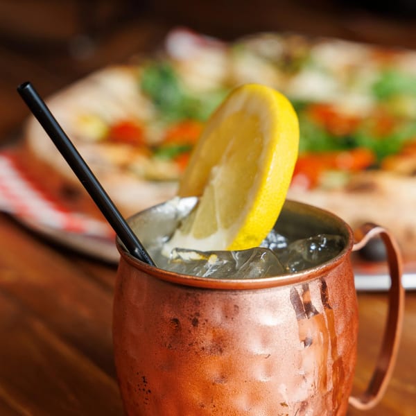 A copper mug with ice, a lemon slice, and a black straw, with a pizza in the background.