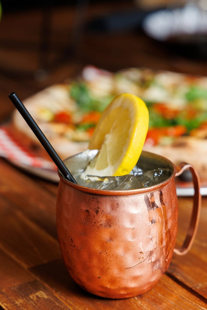 A copper mug with ice, a lemon slice, and a black straw, with a pizza in the background.